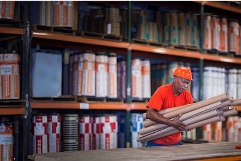 Worker in orange shirt and hard hat carries materials in a Siplast manufacturing facility, showcasing organized product storage and plant tour opportunities.