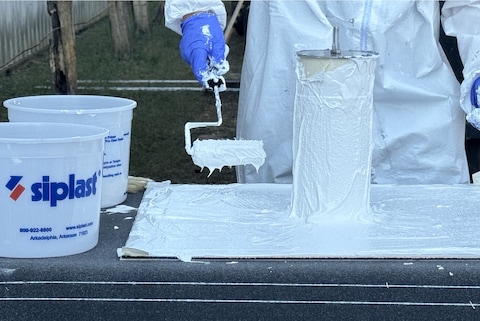 A person wearing protective gear uses a roller to apply a white liquid product to a cylindrical sample, demonstrating a hands-on technical training session for SBS-modified bitumen, PMMA, or other Siplast product applications.