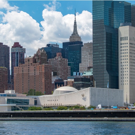 New York City skyline viewed from across the water, featuring the United Nations Headquarters and distant skyscrapers.